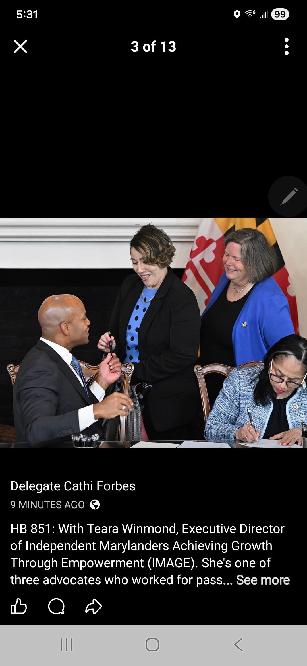 Vier mense glimlag en kommunikeer om 'n lessenaar met dokumente. Twee vroue staan, een hou 'n lêer vas, terwyl 'n man wat voor sit met 'n pen gebaar. Maryland en die VSA se vlae is in die agtergrond sigbaar.