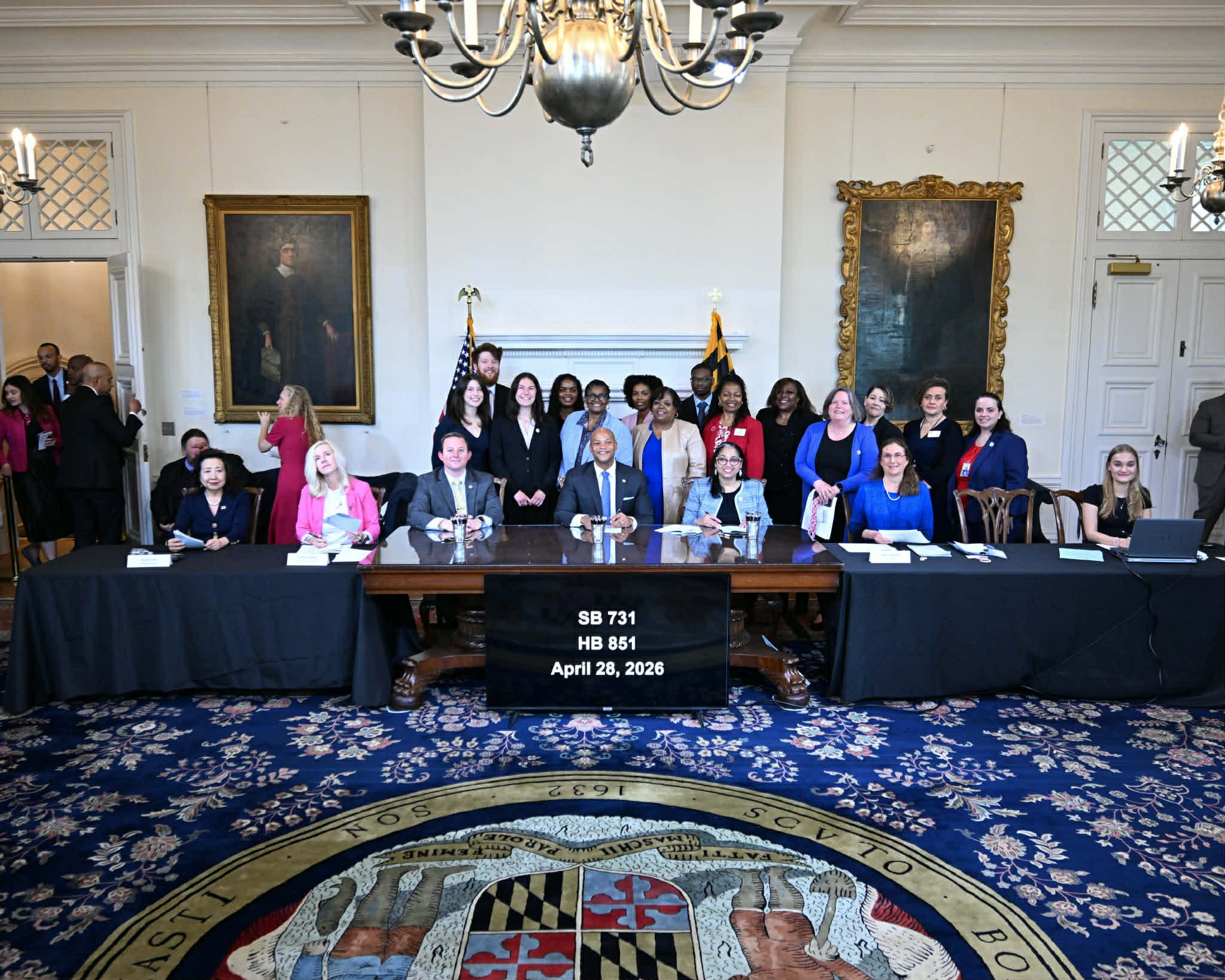 'n Groep mense, insluitend amptenare, poseer binne 'n formele kamer met groot skilderye en vlae. Drie tafels met naamborde lui SB 731, HB 851 en 28 April 2026. Die Maryland-staatseël is op die vloer sigbaar.