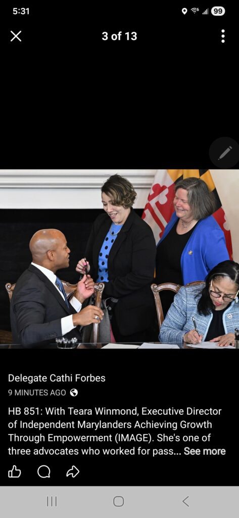 Vier mense glimlag en kommunikeer om 'n lessenaar met dokumente. Twee vroue staan, een hou 'n lêer vas, terwyl 'n man wat voor sit met 'n pen gebaar. Maryland en die VSA se vlae is in die agtergrond sigbaar.